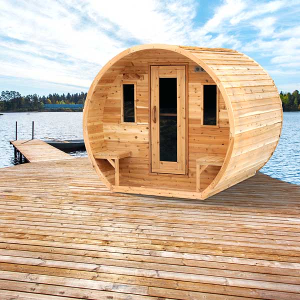 Wooden barrel sauna on a wooden deck by a lake with a clear blue sky.