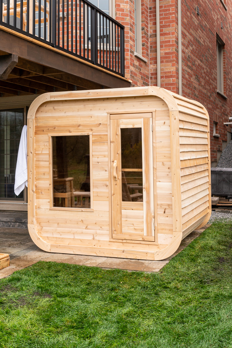 Wooden outdoor cabin with glass doors and windows on a grassy area in front of a brick building.