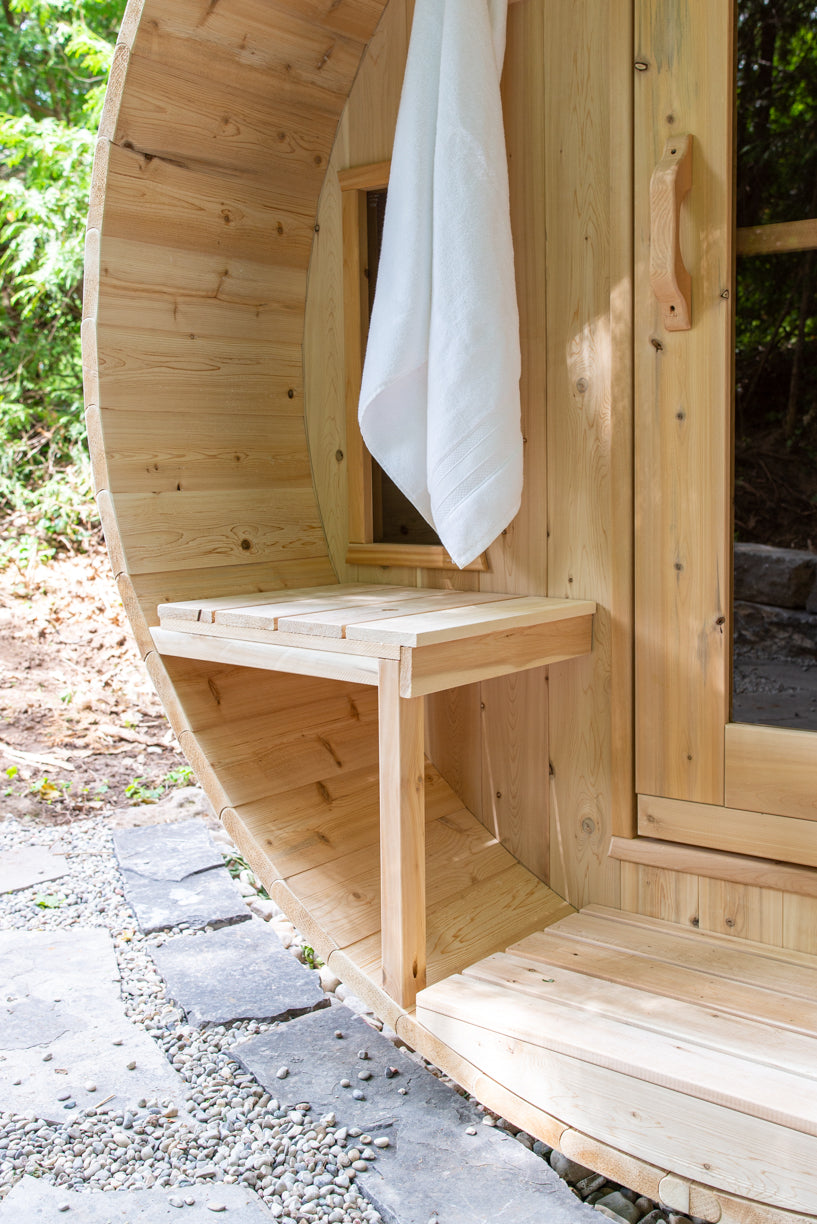 Wooden outdoor barrel sauna cabin with a white towel hanging on porch, surrounded by greenery.