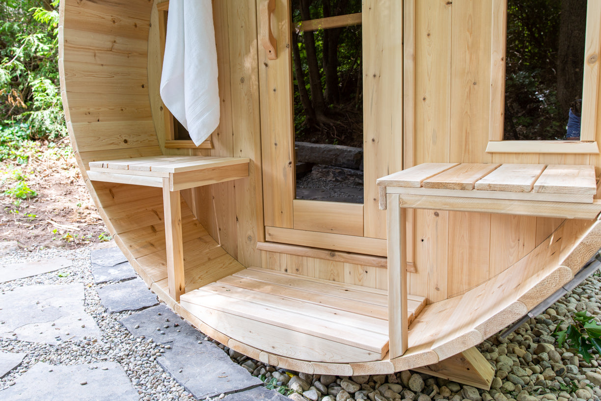 Wooden outdoor sauna with a white towel hanging on the [orch, surrounded by greenery.