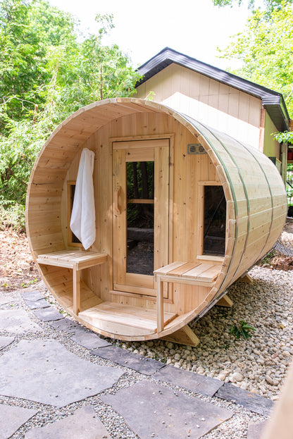 Wooden barrel-shaped sauna in a natural setting with trees and stones.
