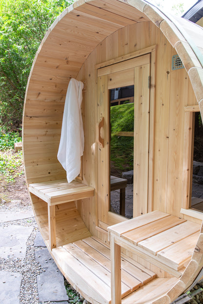 Wooden barrel-shaped sauna with a bench and towel hanging on the porch, surrounded by greenery.