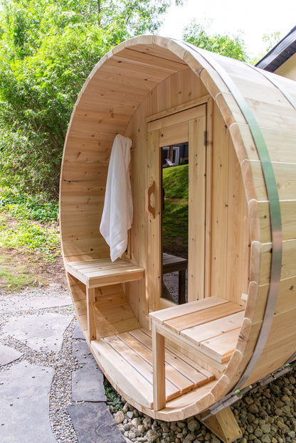 Wooden barrel-shaped outdoor sauna with a white towel hanging on the porch, surrounded by greenery.