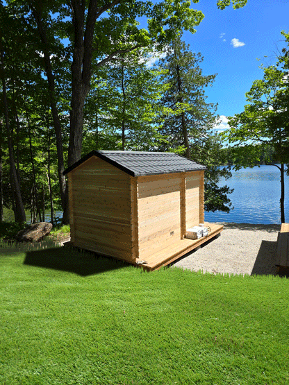 Rear of wooden sauna with changeroom on gravel with lake and trees in the background. 