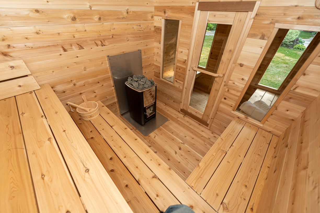 Wooden interior of a cabin with a small stove and windows showing greenery outside.