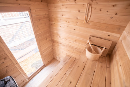 Wooden interior of a sauna with a window and a basket sitting on an upper bench.