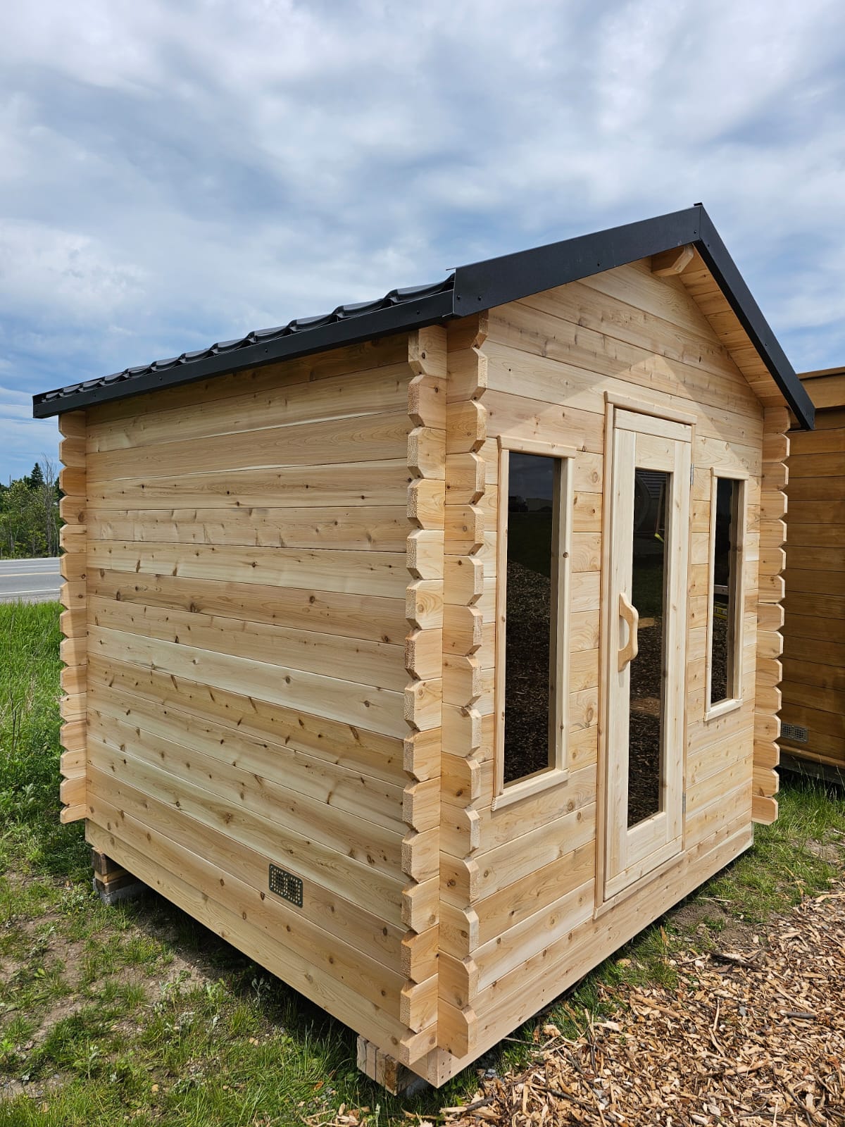Wooden cabin with a black roof on a grassy area with a cloudy sky.