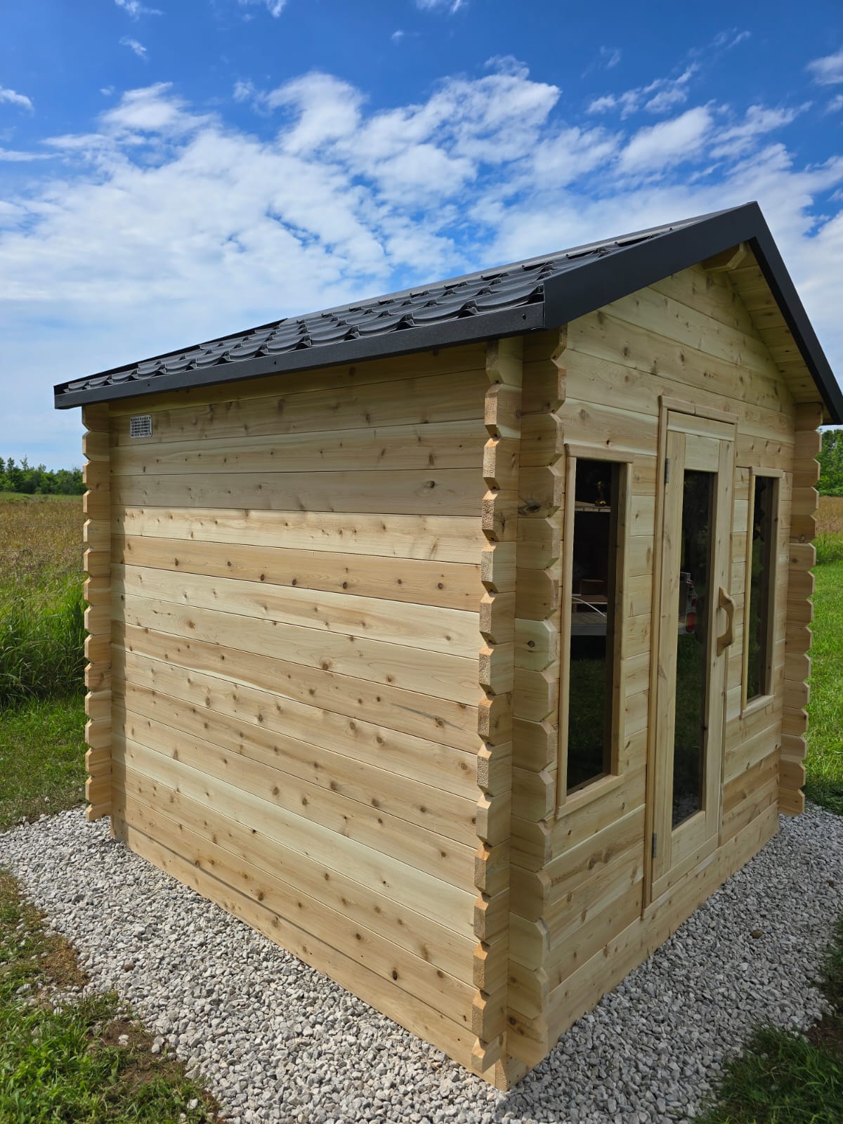 Wooden cabin with a black roof in a grassy area under a blue sky.