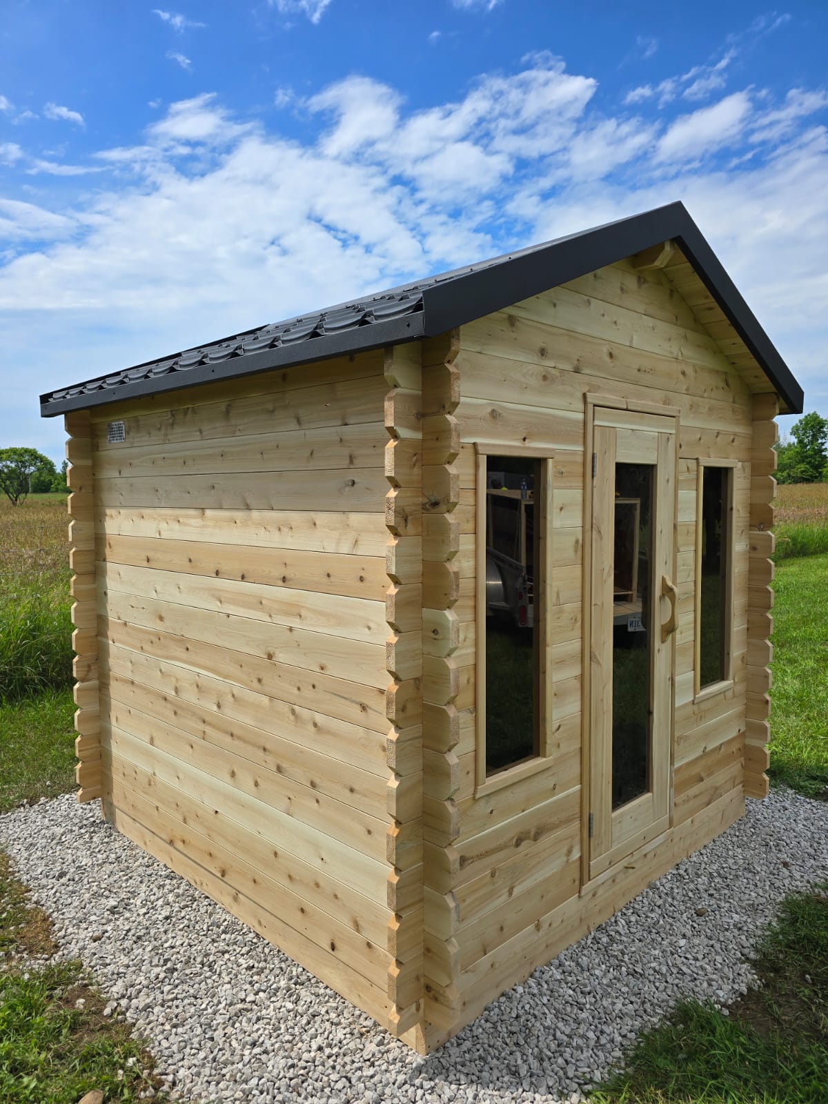 Wooden cabin with a black roof on a gravel foundation against a blue sky.
