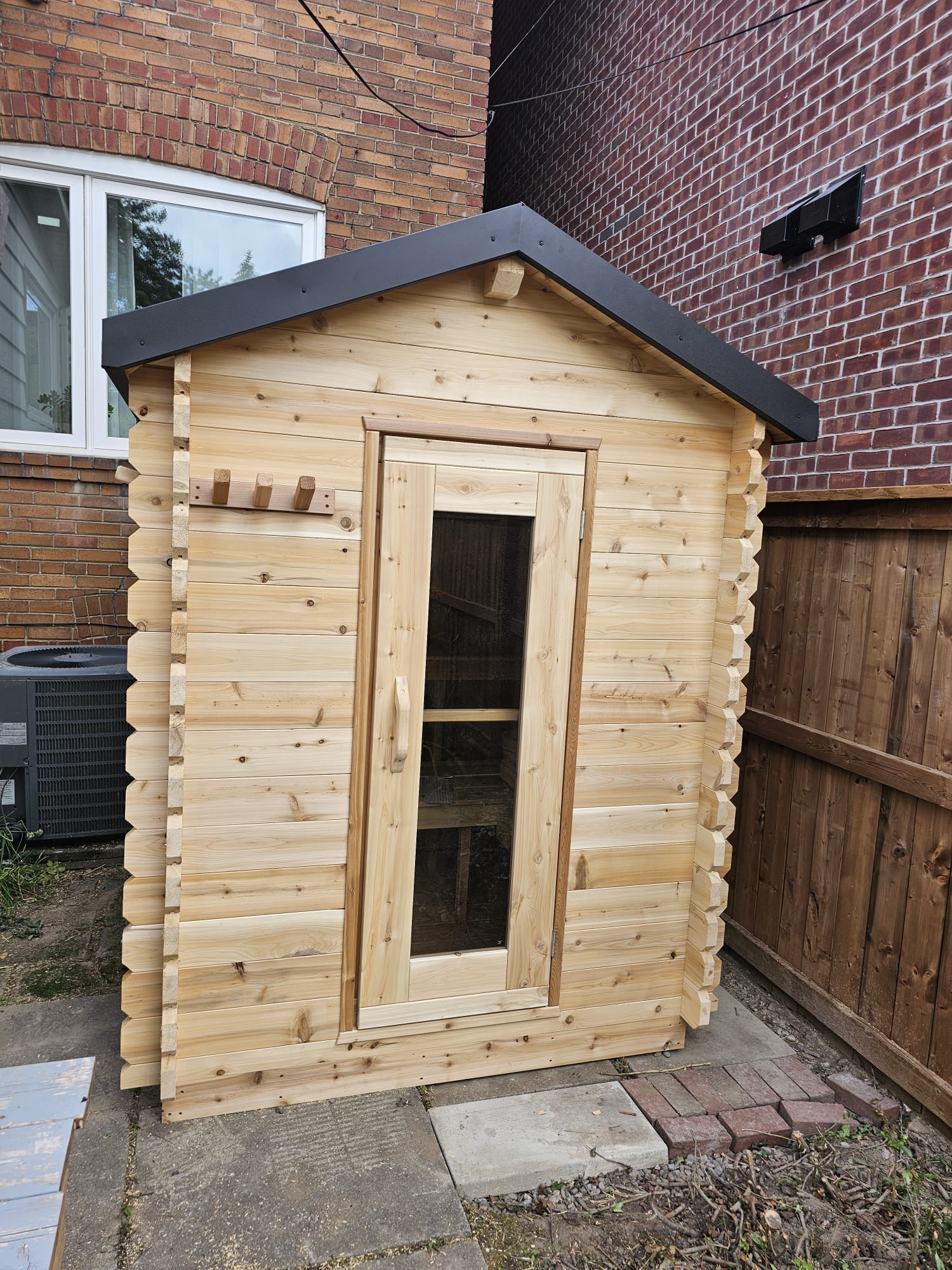 Wooden log cabin shed with a black roof against a brick wall.