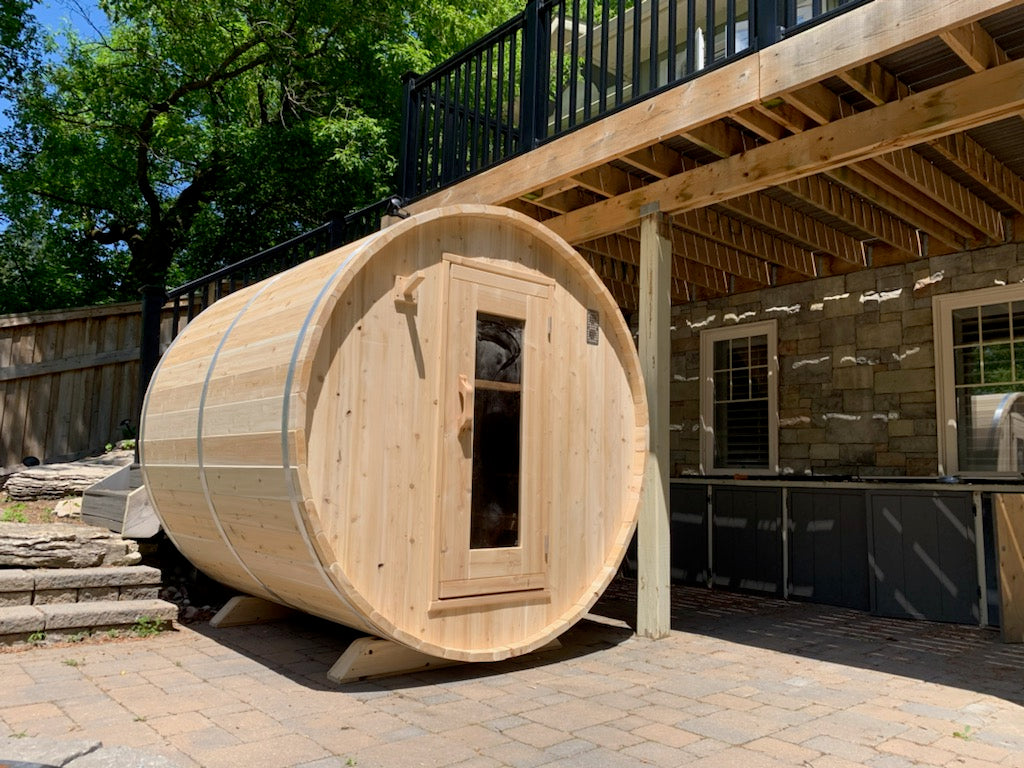 Wooden barrel-shaped sauna on a patio with a wooden deck and trees in the background